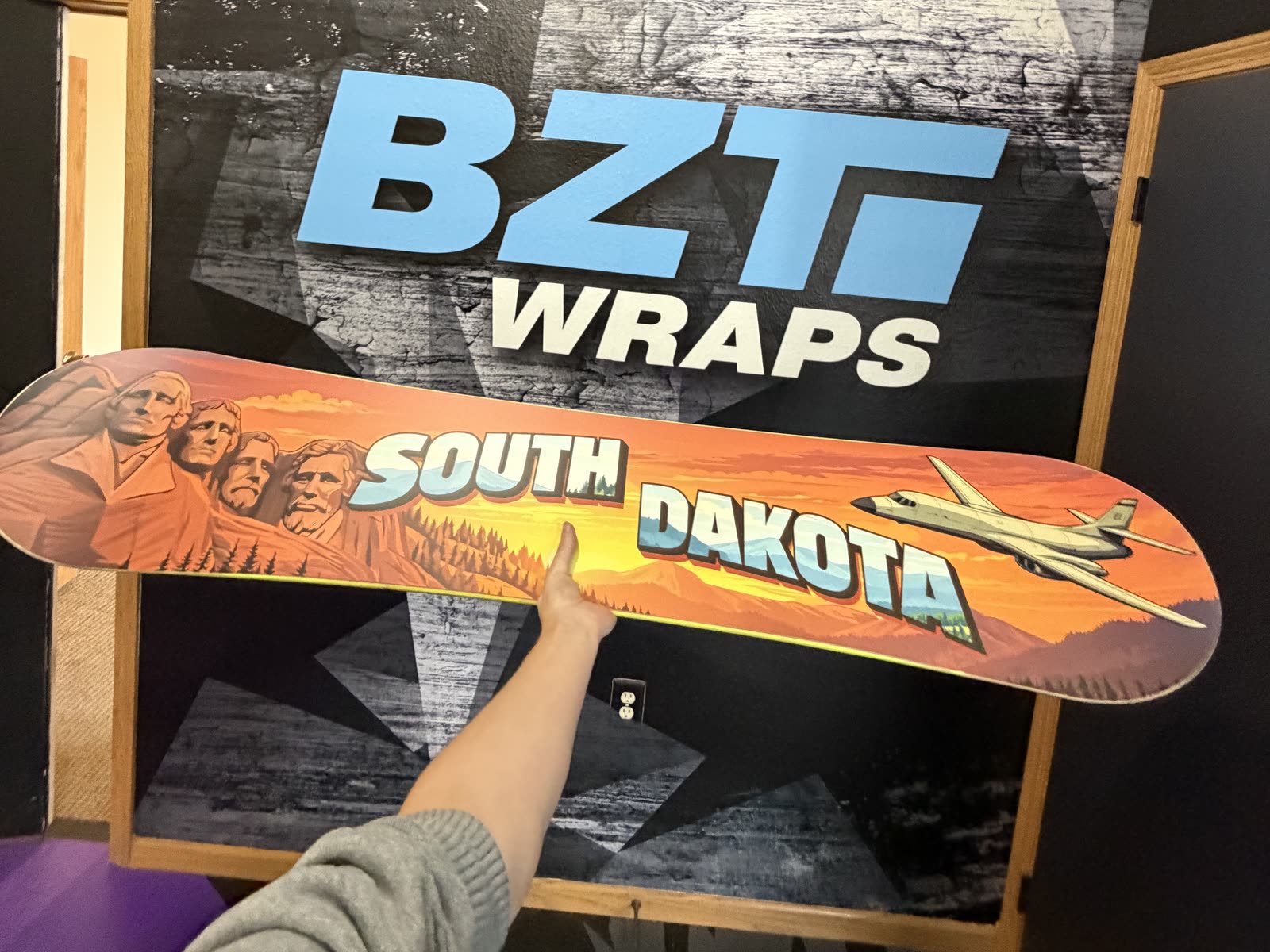 Snowboard with orange South Dakota artwork, Mount Rushmore, and an aircraft, held in front of a BZT Wraps wall graphic.